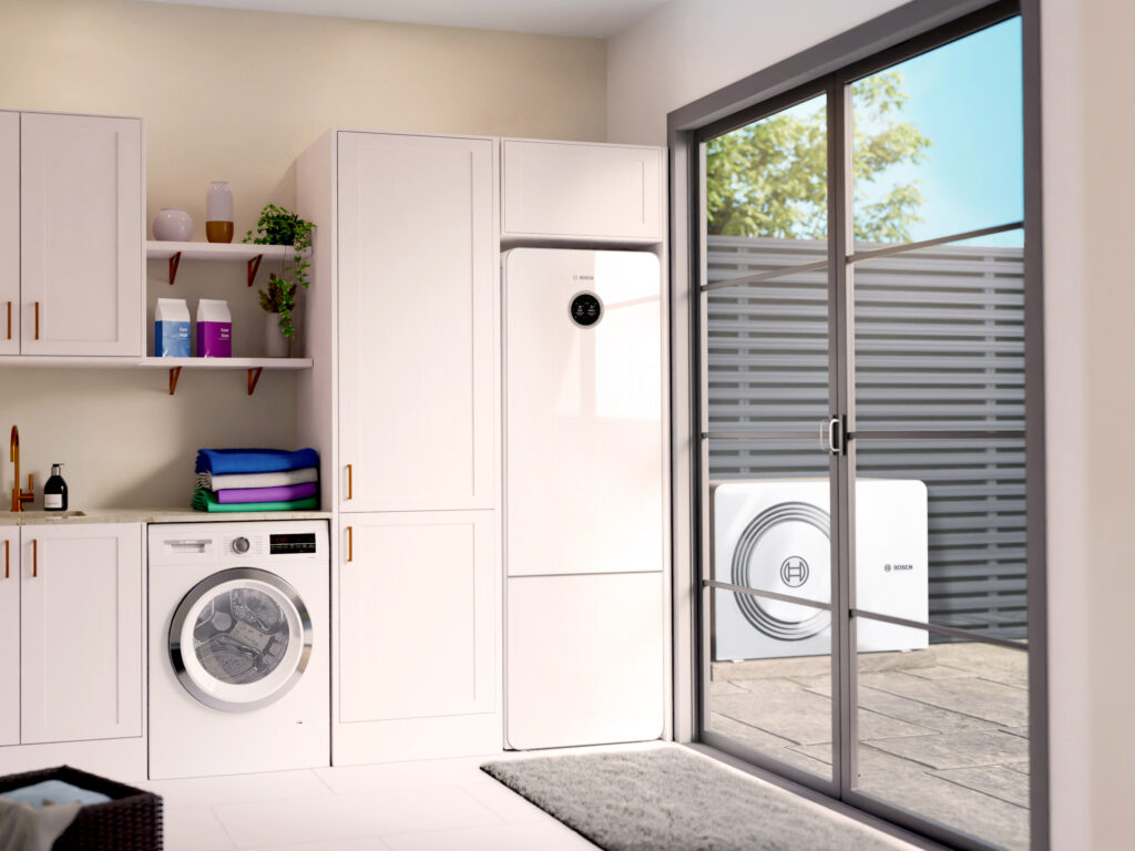 Moderner Hauswirtschaftsraum mit integrierter Bosch Wärmepumpen-Inneneinheit in einem weißen Schrankverbau. Daneben eine Waschmaschine und Regale mit Handtüchern und Waschmittel. Durch die große Glasschiebetür ist die Außeneinheit der Wärmepumpe auf der Terrasse sichtbar.