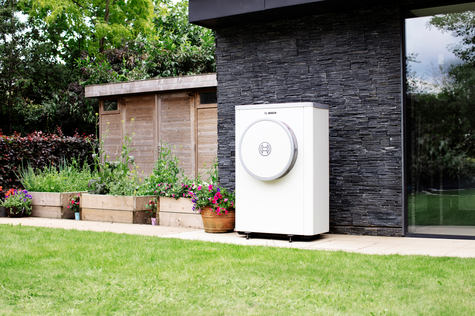 Eine weiße Bosch Wärmepumpe steht an der dunklen Steinwand eines modernen Hauses. Umgeben ist sie von einem gepflegten Garten mit Rasen, Hochbeeten und blühenden Blumen in Töpfen. Rechts spiegelt sich das Grün im Fenster des Hauses. Die Wärmepumpe fügt sich harmonisch in die naturnahe Umgebung ein.