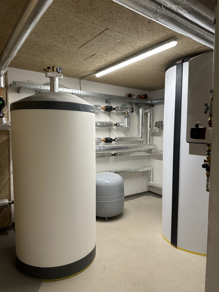 Technikraum mit zwei großen, isolierten Speicher für Heizungs- und Warmwassersysteme. Zahlreiche gedämmte Rohrleitungen verlaufen an den Wänden. Die Anlage ist modern und sauber installiert, unter einer Decke mit integrierter Leuchtstoffröhre.
