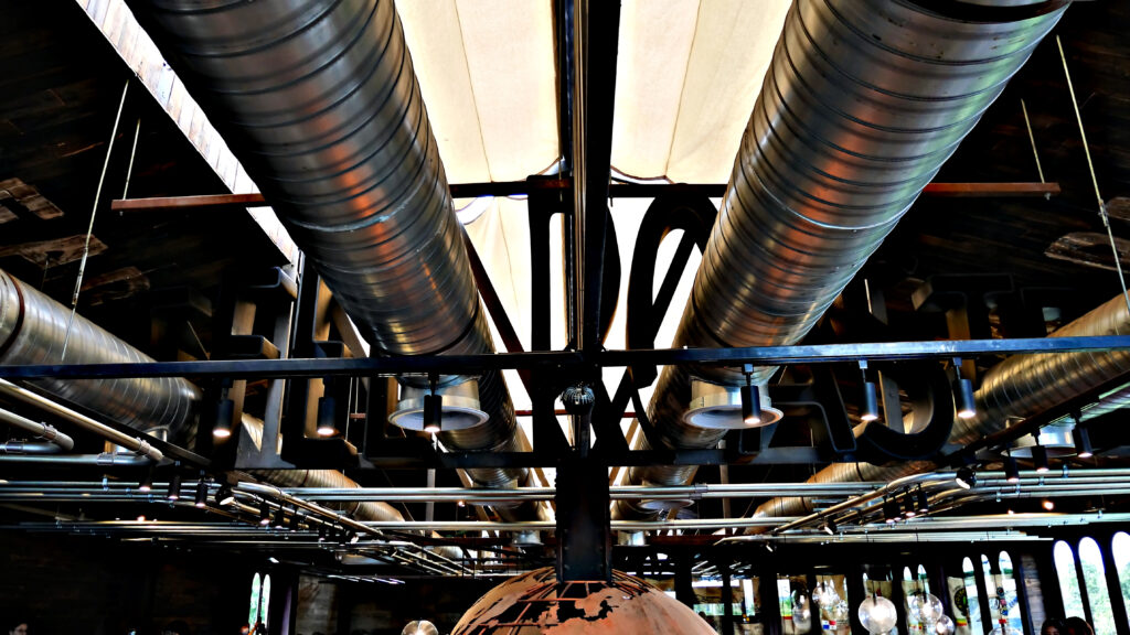 Große, runde Metall-Lüftungsrohre verlaufen unter der Decke eines Gebäudes mit industriellem Design. Sichtbare Leitungen, Rohre und Lampen unterstreichen den modernen, offenen Baustil.