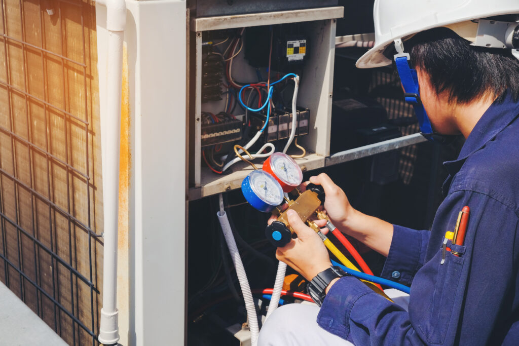 Techniker mit Schutzhelm misst den Druck einer Klimaanlage mit einem Manometer vor geöffnetem Außengerät