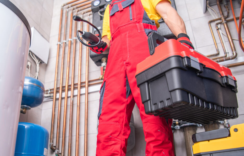 Ein Klempner in roter Arbeitskleidung steht in einem Technikraum vor Rohrleitungen und hält in der rechten Hand einen Werkzeugkasten. In seiner linken Hand hält er ein Gas- oder Lecksuchmessgerät, das verwendet wird, um Gaslecks oder Undichtigkeiten bei Heizungs- oder Rohrsystemen zu erkennen.