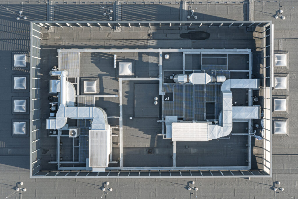 Drohnenaufnahme eines modernen Flachdachs mit umfangreichem Lüftungsanlagensystem. Sichtbar sind zahlreiche Rohre, Schächte und technische Installationen in rechteckiger Anordnung auf dem Dach eines großen Gebäudes.