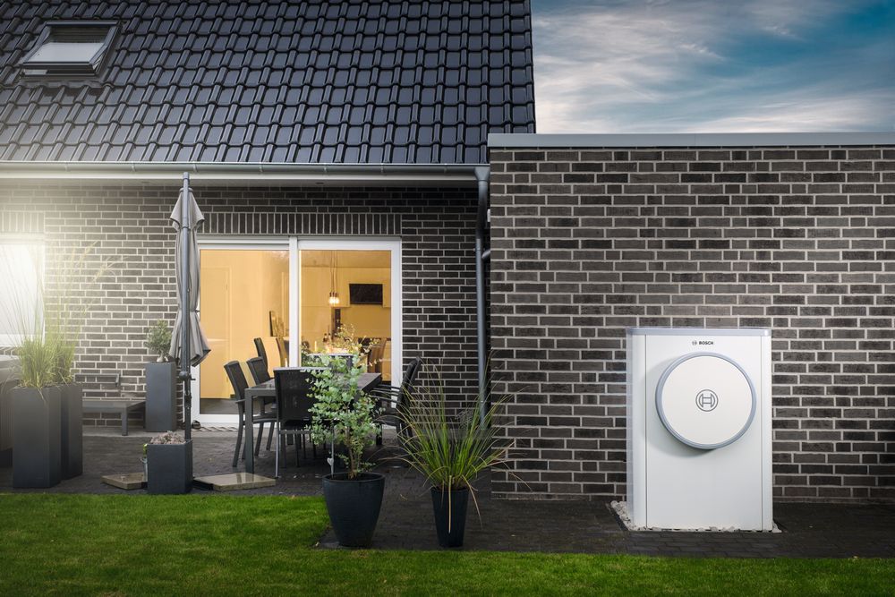 Bosch Wärmepumpe im Außenbereich eines modernen Einfamilienhauses mit dunkler Klinkerfassade. Die kompakte weiße Außeneinheit ist an der Hauswand installiert, umgeben von gepflegtem Garten, Terrasse mit Sitzgruppe und Pflanzen. Die Wärmepumpe dient der umweltfreundlichen Heiz- und Kühltechnik.
