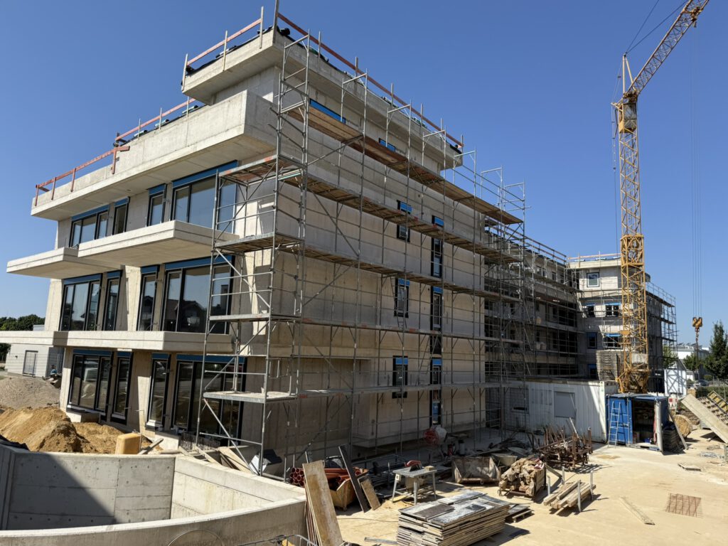 Ein modernes Mehrfamilienhaus befindet sich im Rohbau. Das Gebäude ist eingerüstet, ein gelber Baukran ragt in den blauen Himmel. Auf der Baustelle liegen Baumaterialien und Werkzeuge, während im Vordergrund ein bereits betonierter Bereich erkennbar ist. Der Rohbau zeigt große Fensteröffnungen und eine klare, kantige Architektur.