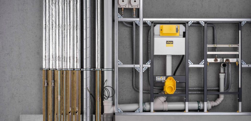 Blick auf eine geöffnete Vorwandinstallation mit sichtbaren Sanitär- und Rohrsystemen. Zu sehen sind Wasserleitungen, ein Viega-Spülkasten, Abflussrohre, Kupfer- und Verbundrohre sowie eine stabile Metallrahmenkonstruktion für die spätere Wandverkleidung.