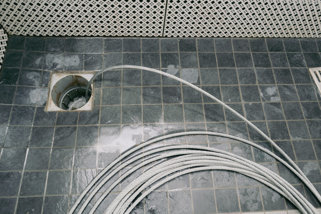 Eine Rohrreinigungsspirale wird in einen Bodenabfluss eingeführt, um eine Verstopfung in der Abwasserleitung zu beseitigen. Der Abfluss befindet sich in einem gefliesten Raum mit dunklen Bodenfliesen.