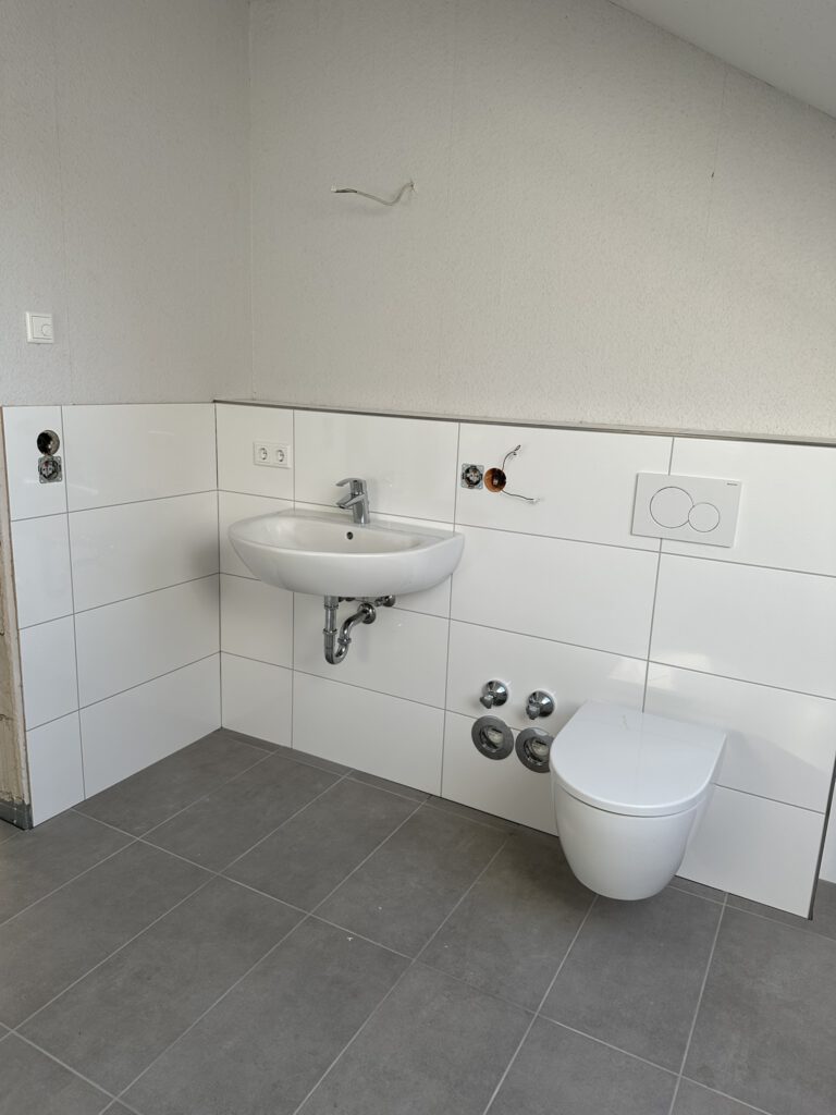 Teilweise eingerichtetes Badezimmer mit weiß gefliestem Wandbereich, einem montierten Wand-WC und einem Waschbecken mit sichtbarem Siphon. Elektro- und Wasseranschlüsse sind vorbereitet, einige Installationen wie Spiegel und Armaturen fehlen noch.