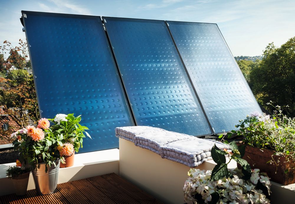 Drei blaue Solarkollektoren auf einer sonnigen Dachterrasse, umgeben von bunten Blumen in Töpfen und einer Sitzbank mit Polster. Im Hintergrund sind Bäume und Häuser zu sehen.
