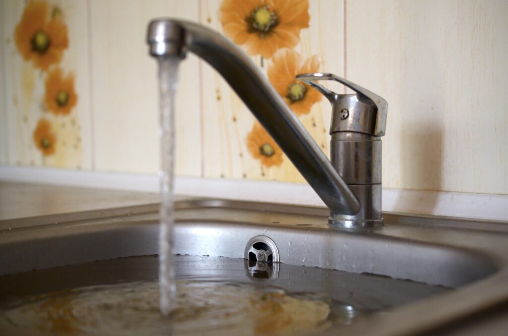 Aus einem Wasserhahn läuft Wasser in ein verstopftes Spülbecken, das sich langsam füllt. Im Hintergrund ist ein Fliesenspiegel mit orangefarbenen Blumen im Retro-Stil zu sehen. Das Wasser staut sich sichtbar im Becken.