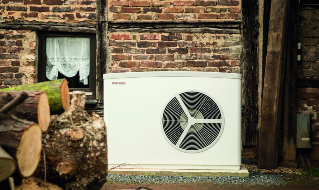 Eine moderne Außeneinheit einer Wärmepumpe der Marke Stiebel Eltron steht vor der Backsteinwand eines alten Fachwerkhauses. Im Vordergrund liegt gestapeltes Brennholz, im Hintergrund ein kleines Fenster mit Gardine. Die Szene zeigt einen Kontrast zwischen moderner Heiztechnik und traditioneller Bauweise.